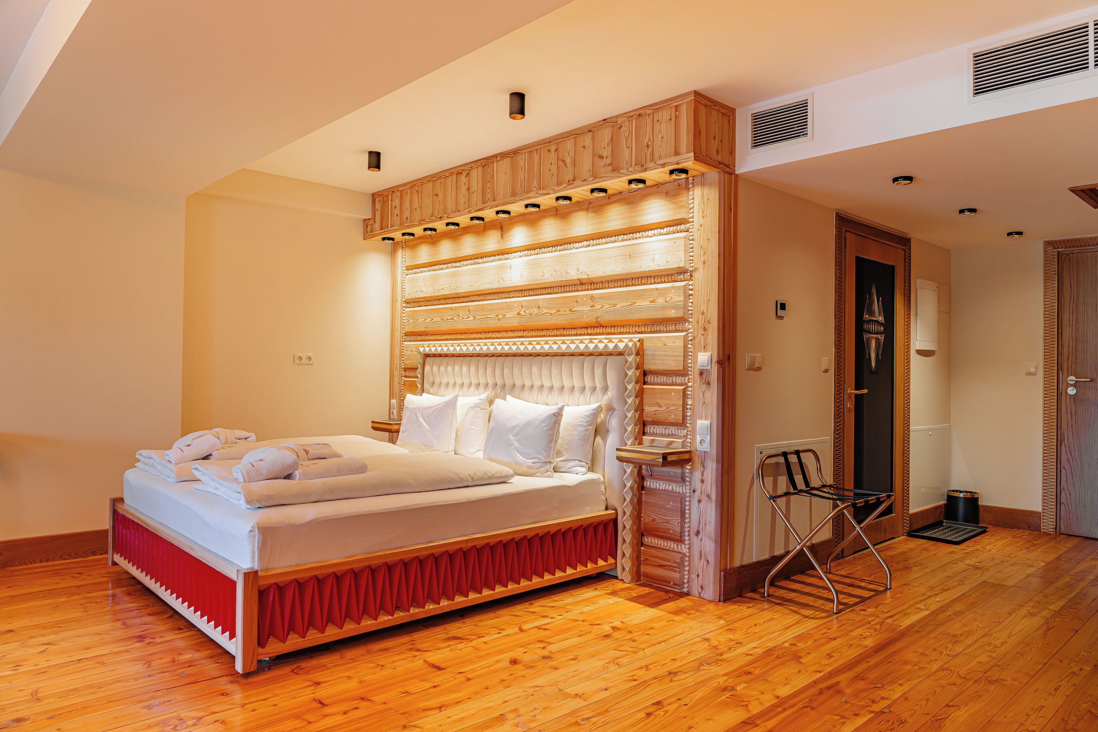 A bedroom with a wooden headboard, a bed with white linens, a red accent on the bed frame, a small table, and a chair.
