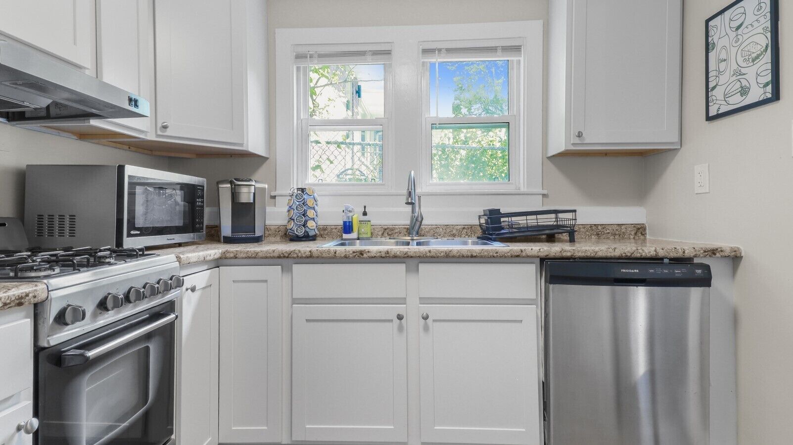 Una cocina con armarios blancos, lavavajillas de acero inoxidable, microondas, estufa y ventana con vista a la vegetación.