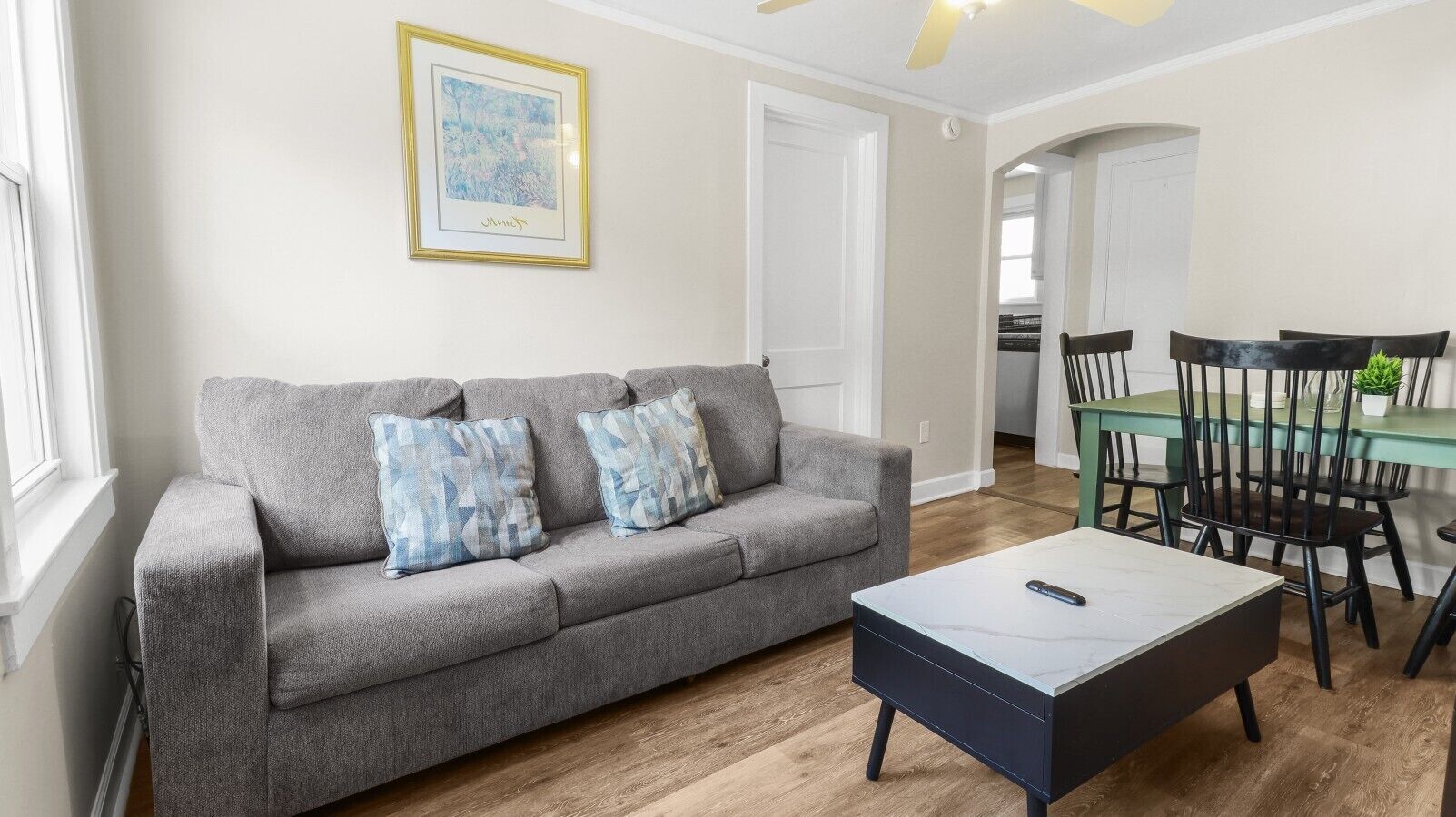 A living room with a grey sofa, two patterned cushions, a marble coffee table, and a dining area with a green table and black chairs.