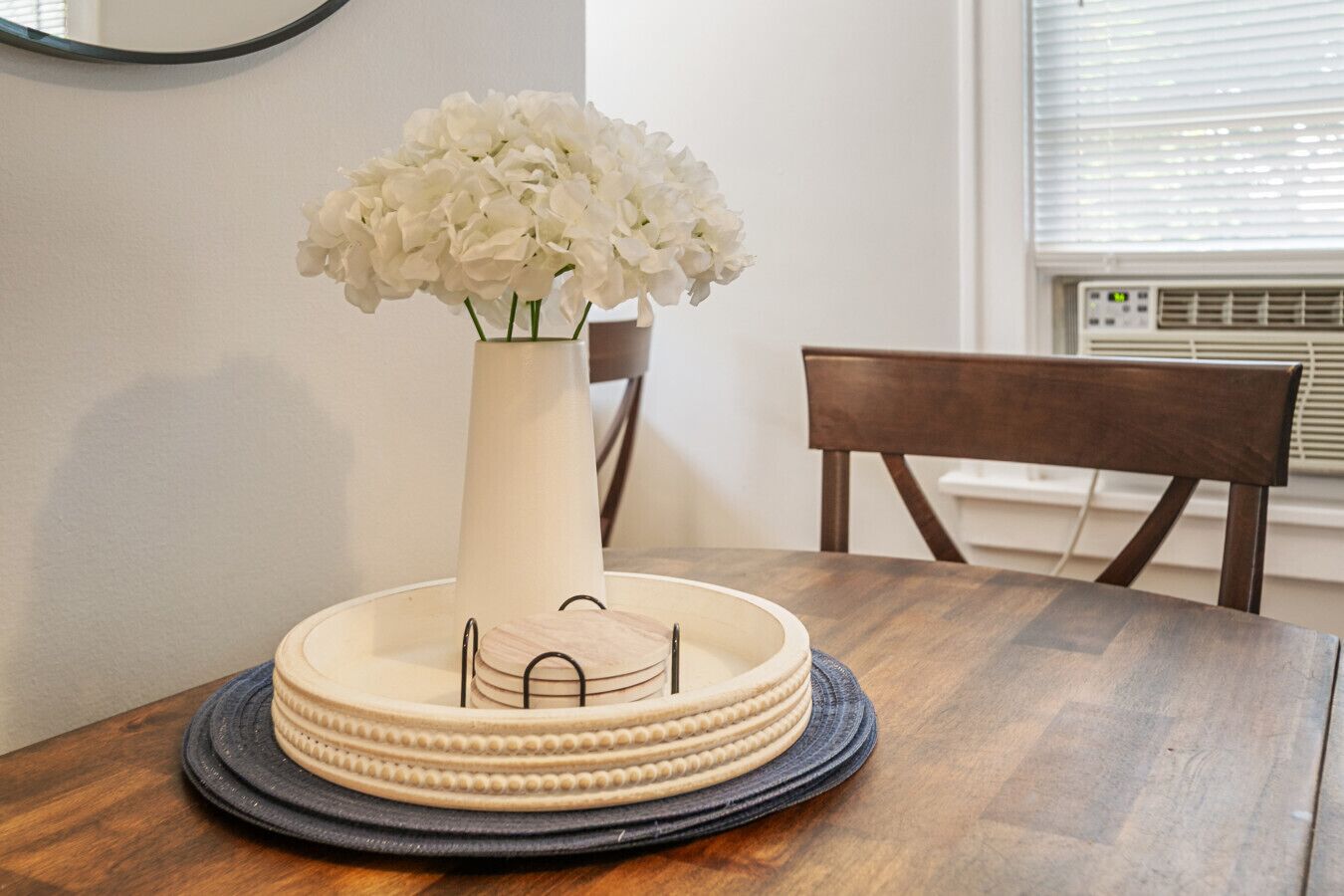 Une table à manger ornée d’un vase de fleurs blanches, d’une assiette décorative et d’une chaise.