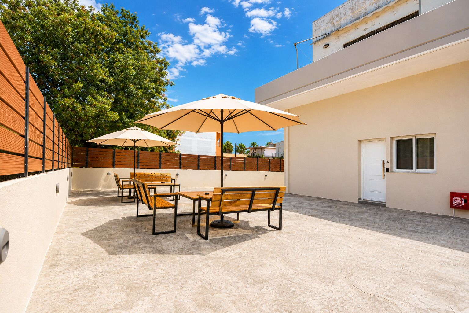 Un patio exterior con mobiliario de madera y sombrillas, una puerta blanca y vista a un edificio al fondo.