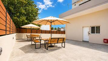 Un patio exterior con mobiliario de madera y sombrillas, una puerta blanca y vista a un edificio al fondo.