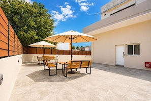 An outdoor patio with wooden furniture and umbrellas, a white door, and a view of a building in the background.