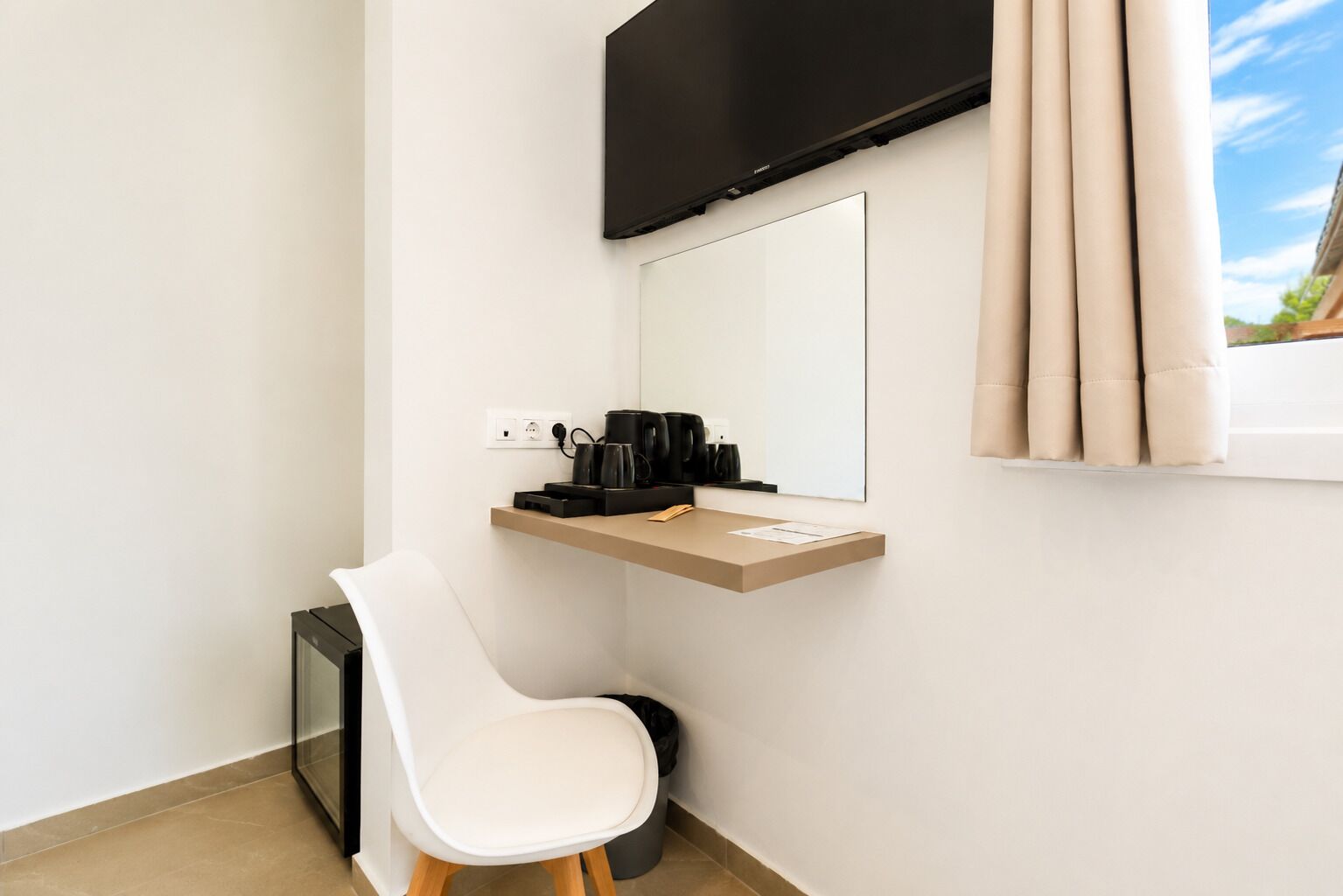 Una habitación de hotel moderna con una televisión de pantalla plana montada en la pared, un pequeño escritorio con cafetera, una silla blanca y una ventana con cortinas.