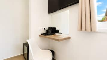 Una habitación de hotel moderna con una televisión de pantalla plana montada en la pared, un pequeño escritorio con cafetera, una silla blanca y una ventana con cortinas.