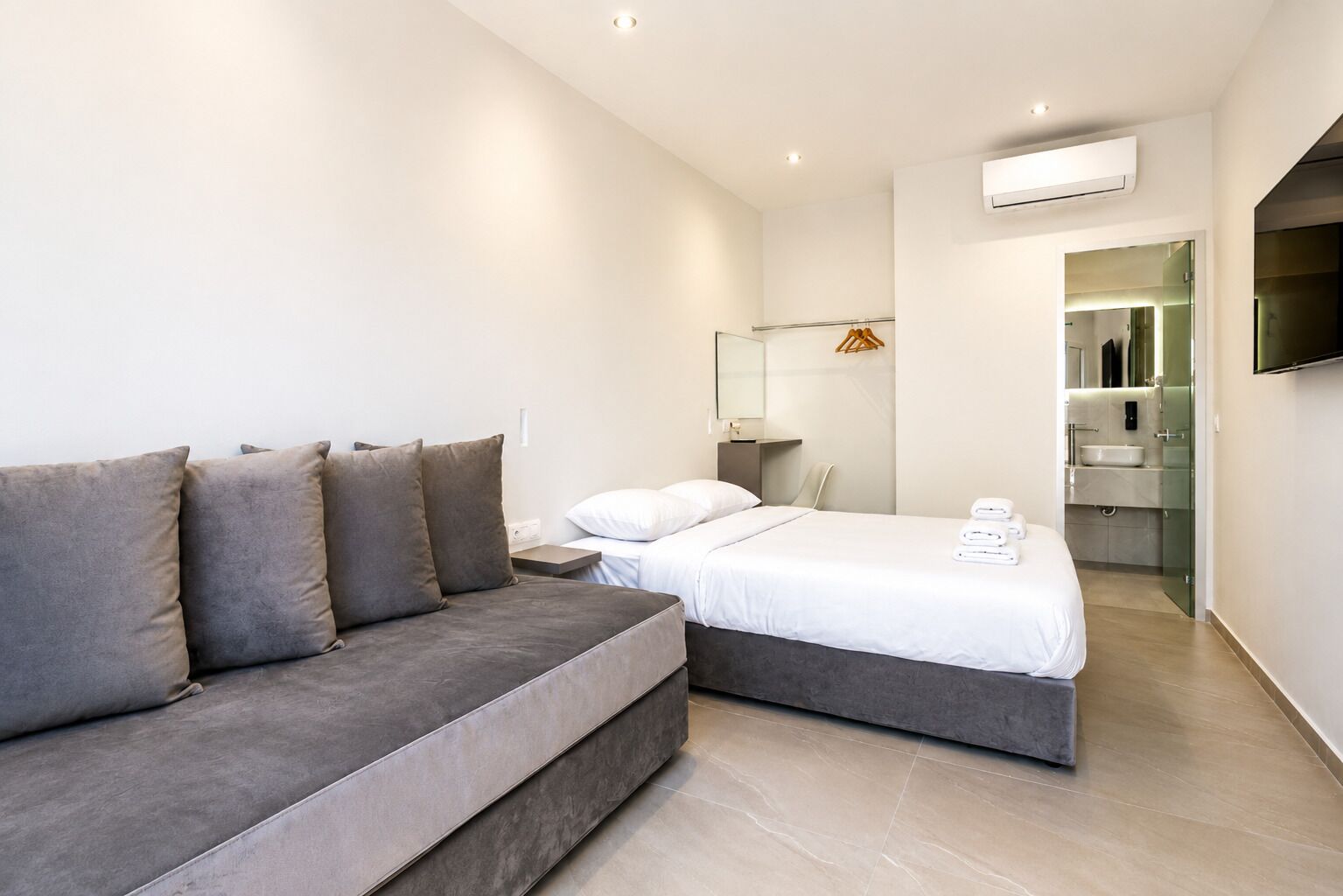 Ein modernes Hotelzimmer mit grauem Sofa, einem Bett mit weißen Leinen, einem kleinen Nachttisch, einem Spiegel und einem an der Wand montierten Fernseher.