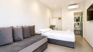 Ein modernes Hotelzimmer mit grauem Sofa, einem Bett mit weißen Leinen, einem kleinen Nachttisch, einem Spiegel und einem an der Wand montierten Fernseher.