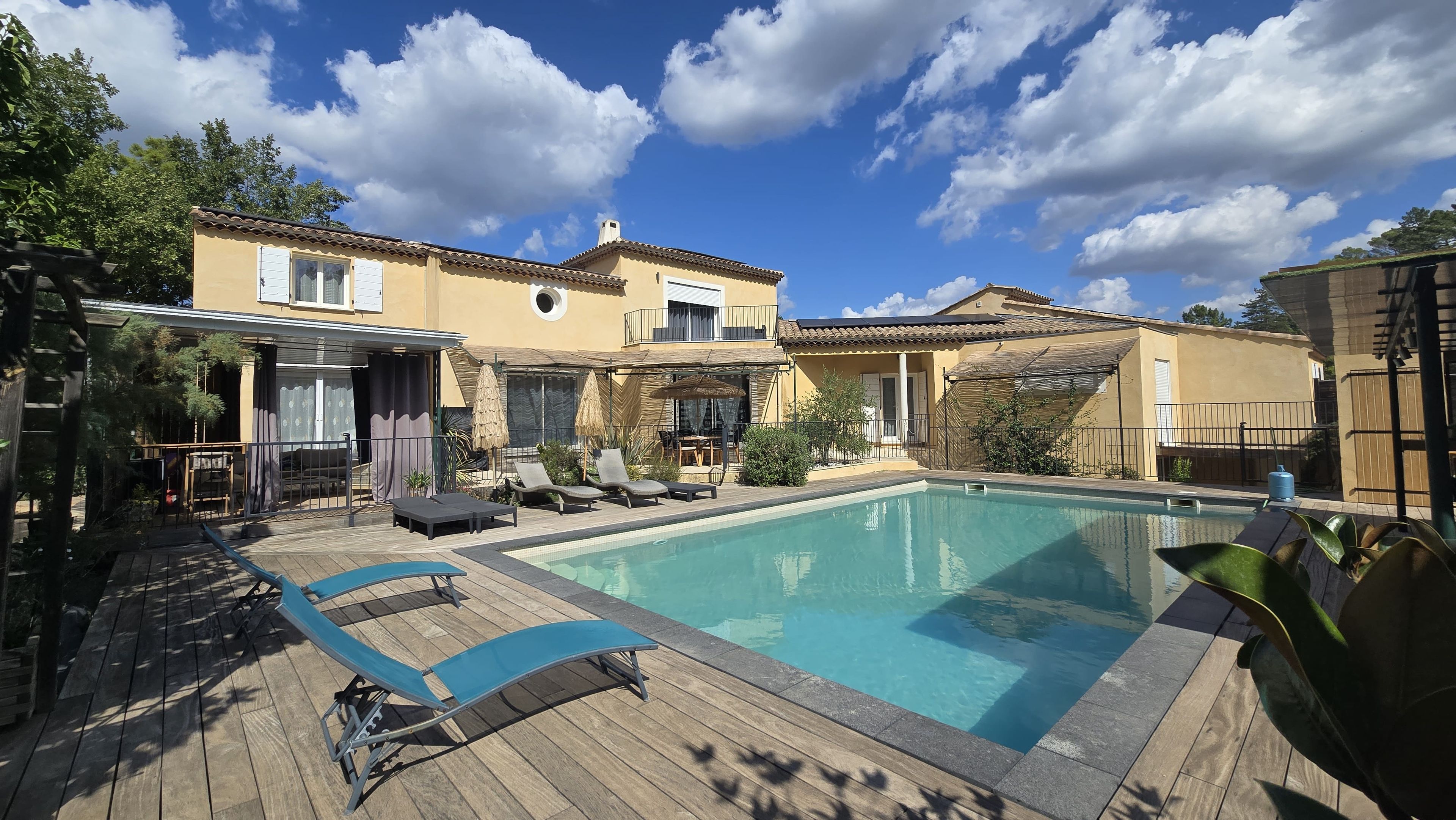A poolside area with lounge chairs and a pool, surrounded by a house with a tiled roof and a covered patio.