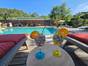 Dos vasos de cóctel sobre una mesa de madera junto a una piscina con sillones de descanso y una cubierta de madera.