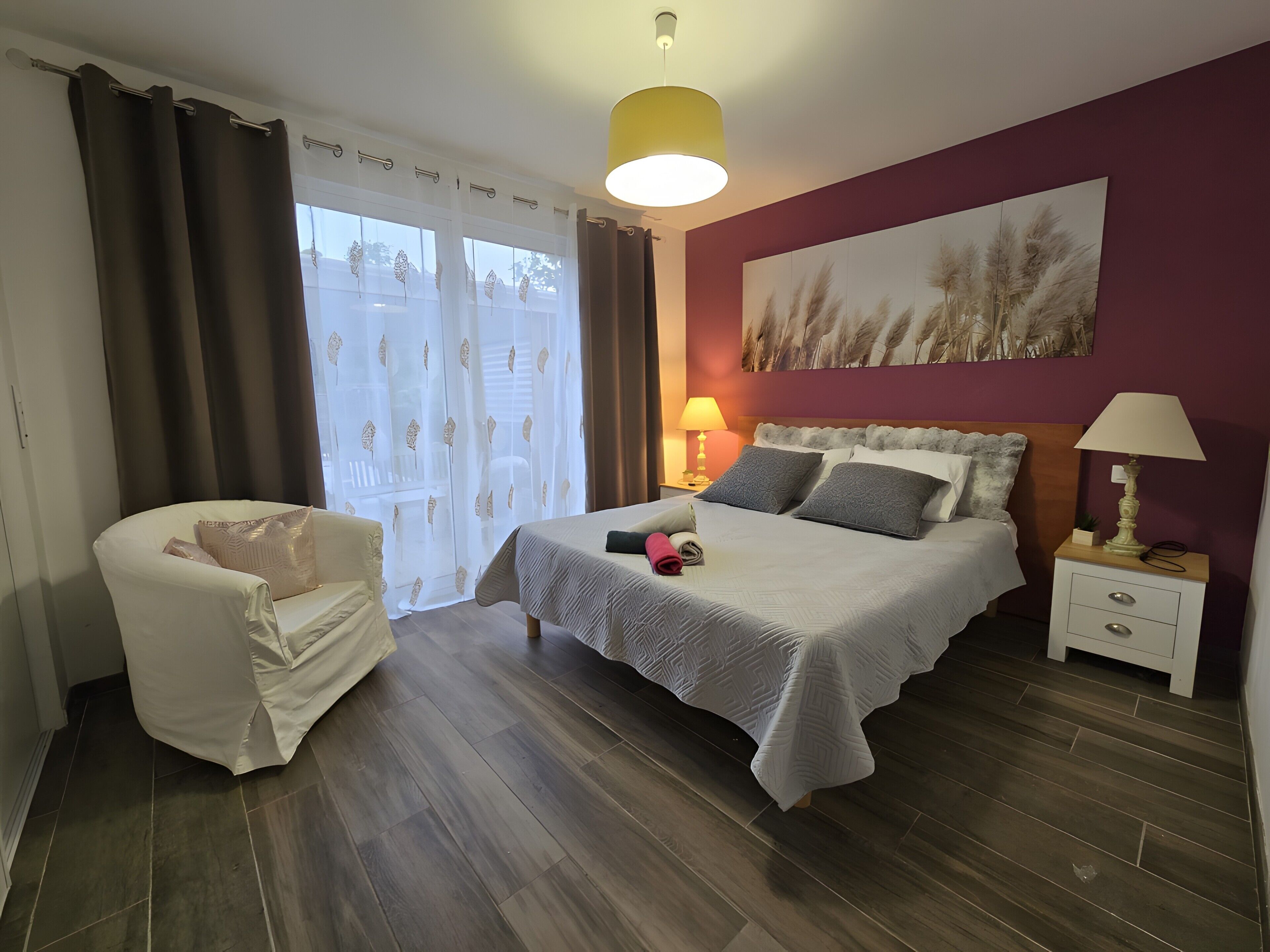 Un dormitorio con una cama, una silla, una mesita de noche, una lámpara y un cuadro en la pared.