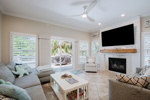Ein Wohnzimmer mit Sofa, Sessel, Couchtisch, Kamin, Fernseher und großen Fenstern mit Jalousien.