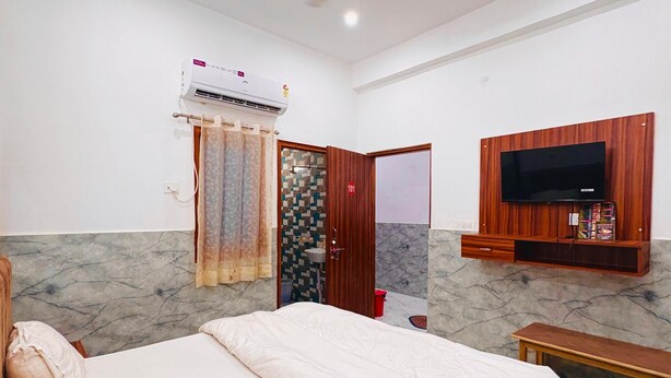 A bedroom with a bed, ceiling fan, air conditioning unit, TV, and a bathroom visible through an open door. - Hotel Vrinda Nikunj Dham (Vrindavan)