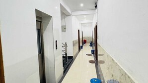 A corridor with white walls, a tiled floor, and a blue bucket on the right. - Hotel Vrinda Nikunj Dham (Vrindavan)