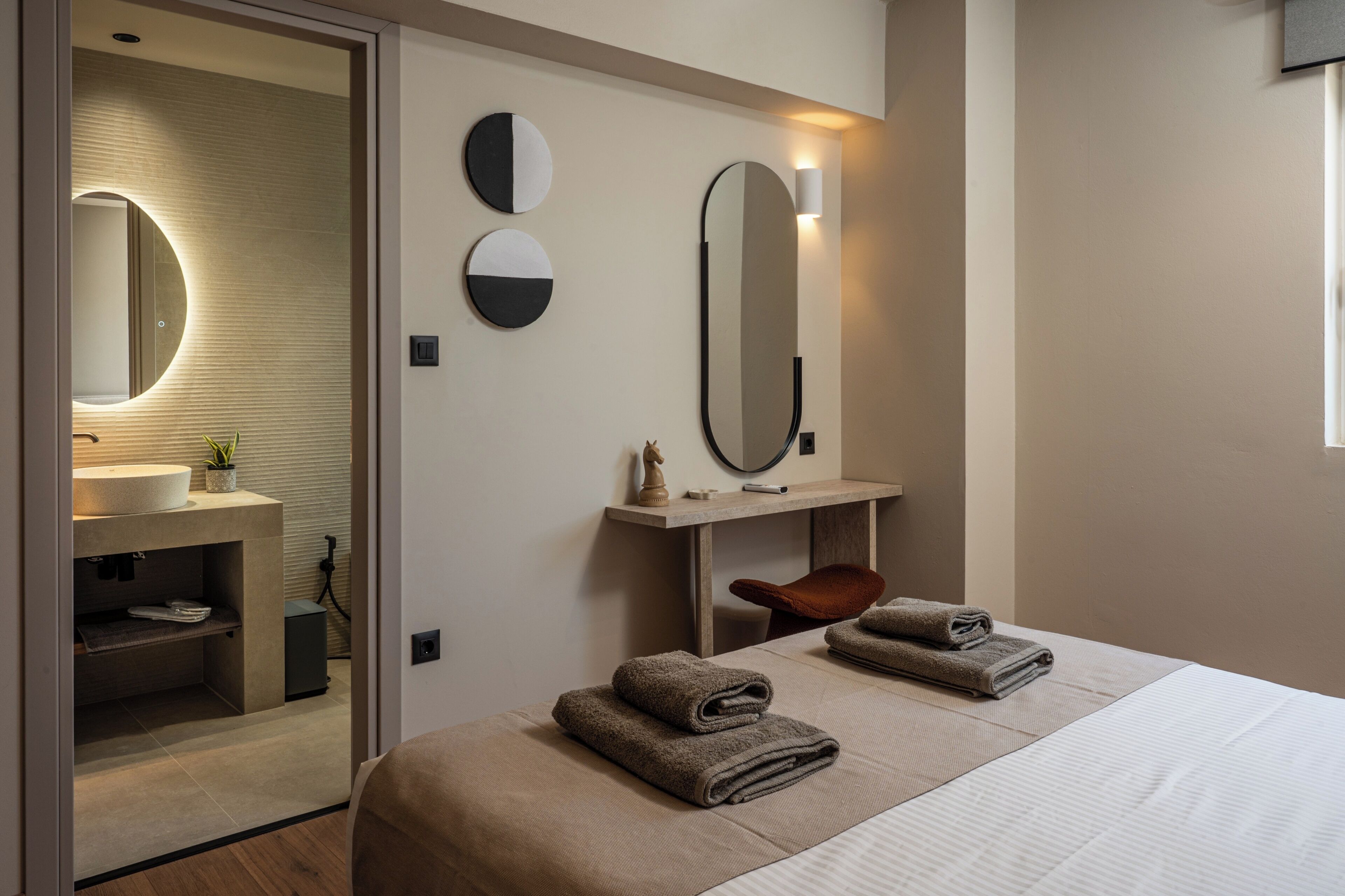 Habitación de hotel moderna con cama, dos espejos en la pared, una mesita de noche, un escritorio pequeño y un baño visible a través de una puerta abierta.
