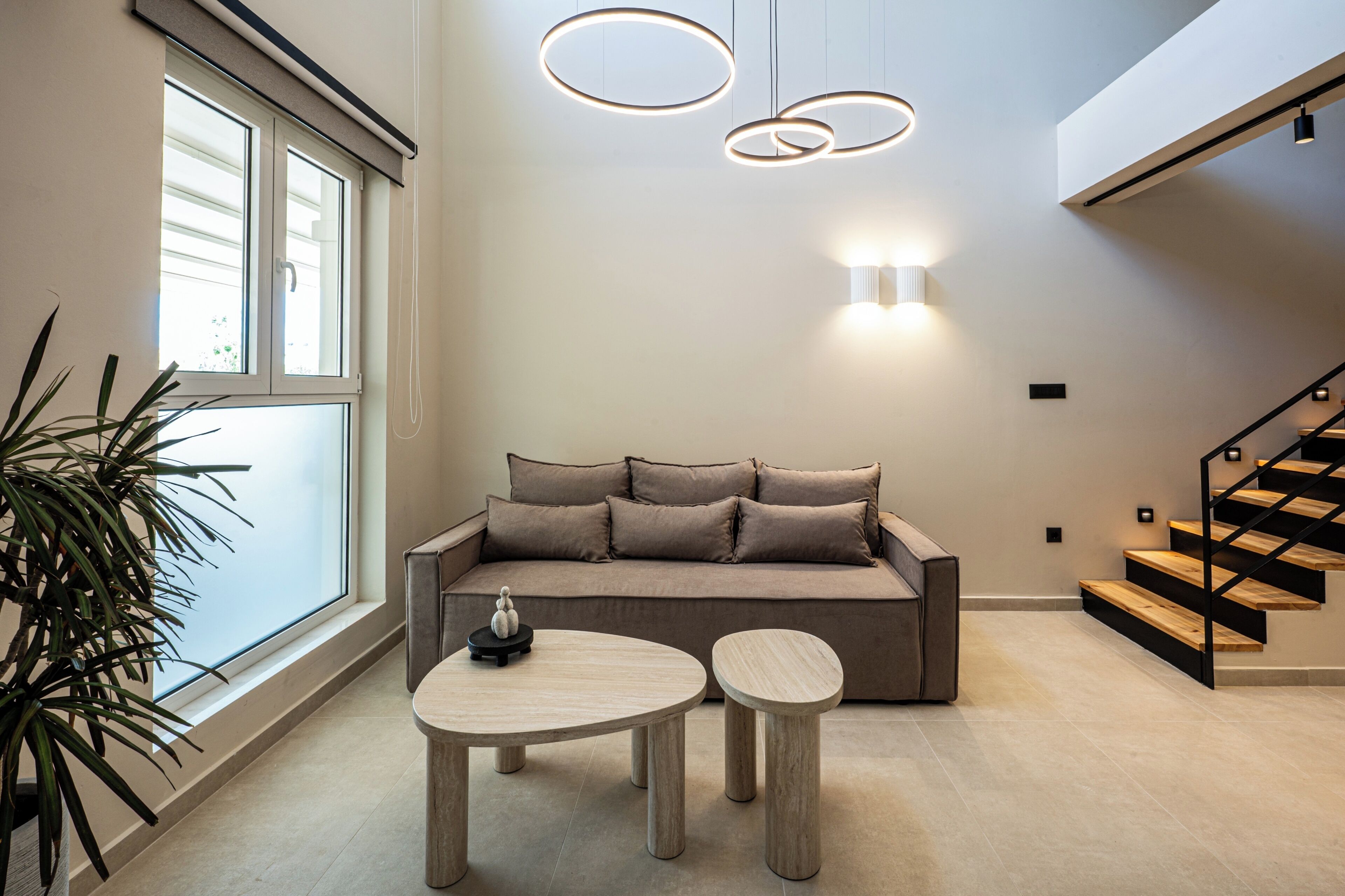 A modern living room with a sofa, two round tables, a staircase, and a large window.