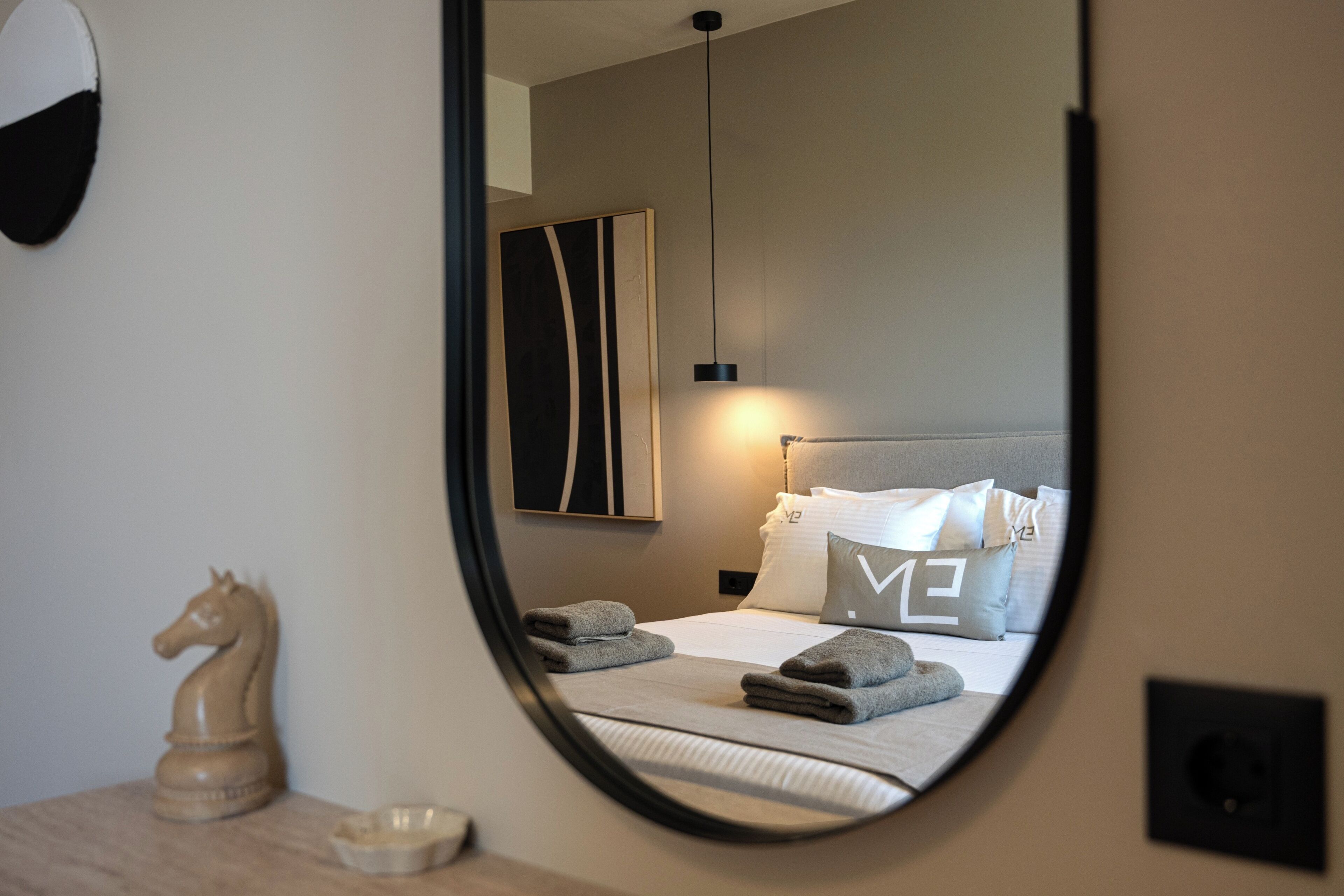 Ein modernes Hotelzimmer mit einem runden Spiegel, der ein ordentlich bezogenes Bett, ein an Wand befestigtes Kunstwerk und eine kleine Holzpferd-Dekoration widerspiegelt.