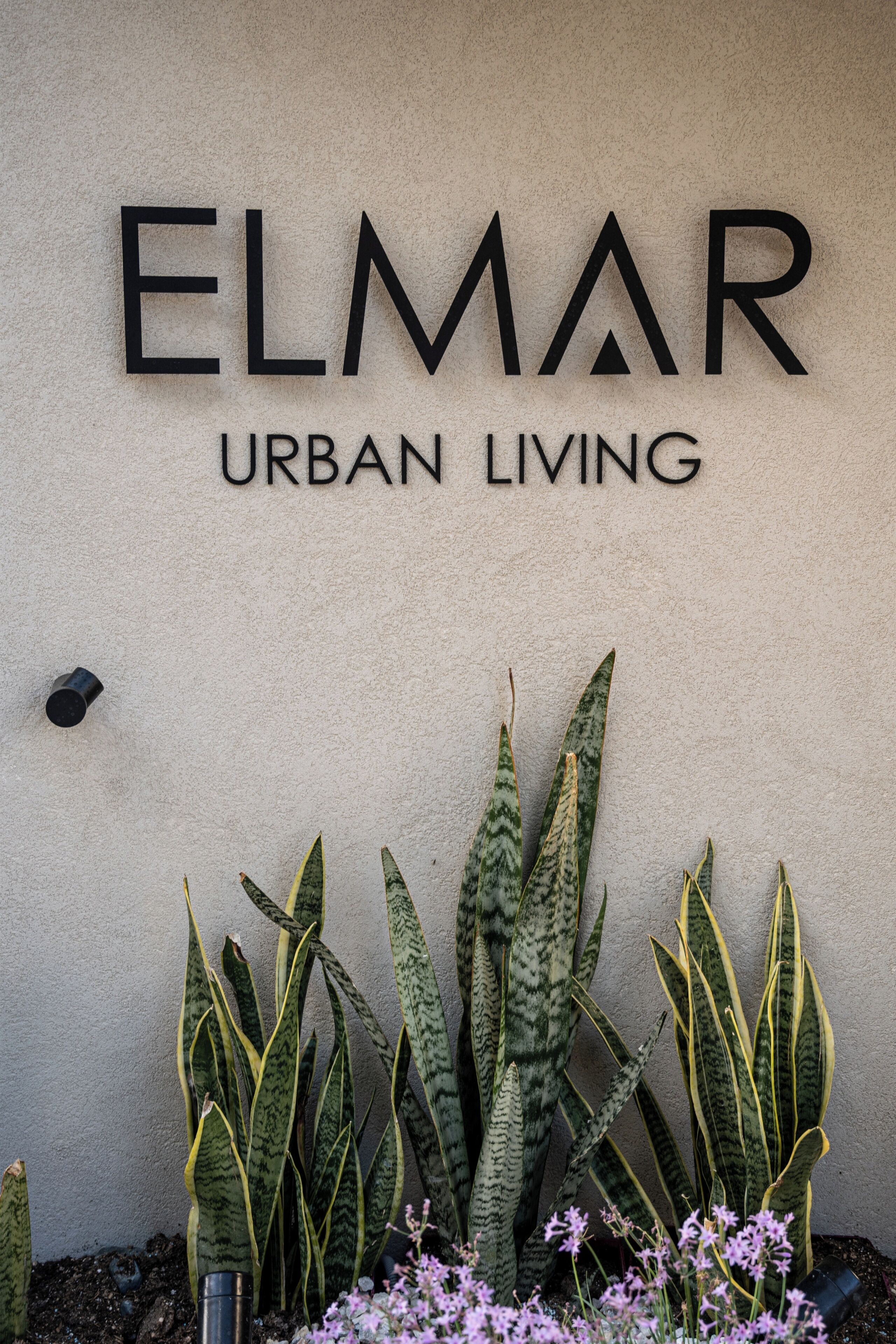 En skylt för ”ELMAR URBAN LIVING” med en vägg i strukturerad yta som bakgrund och dekorativa växter.