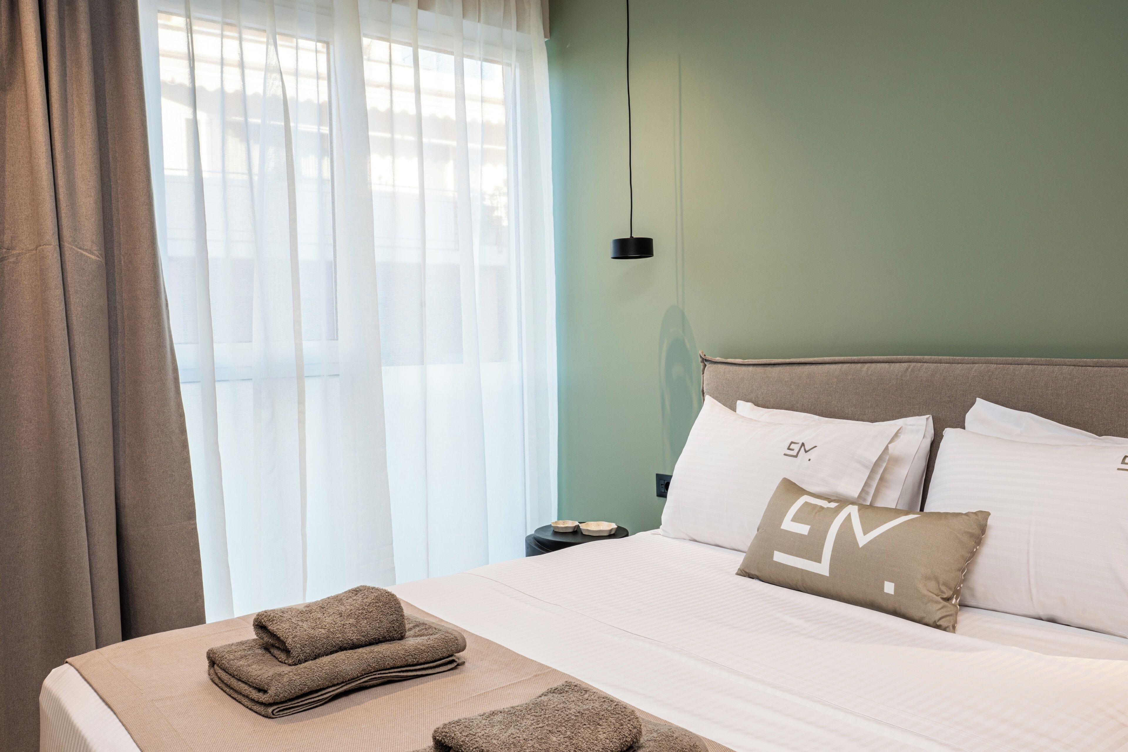 Un letto rifatto con cura, una coperta beige e asciugamani, un cuscino con un logo, un comodino con una lampada e tende leggere.