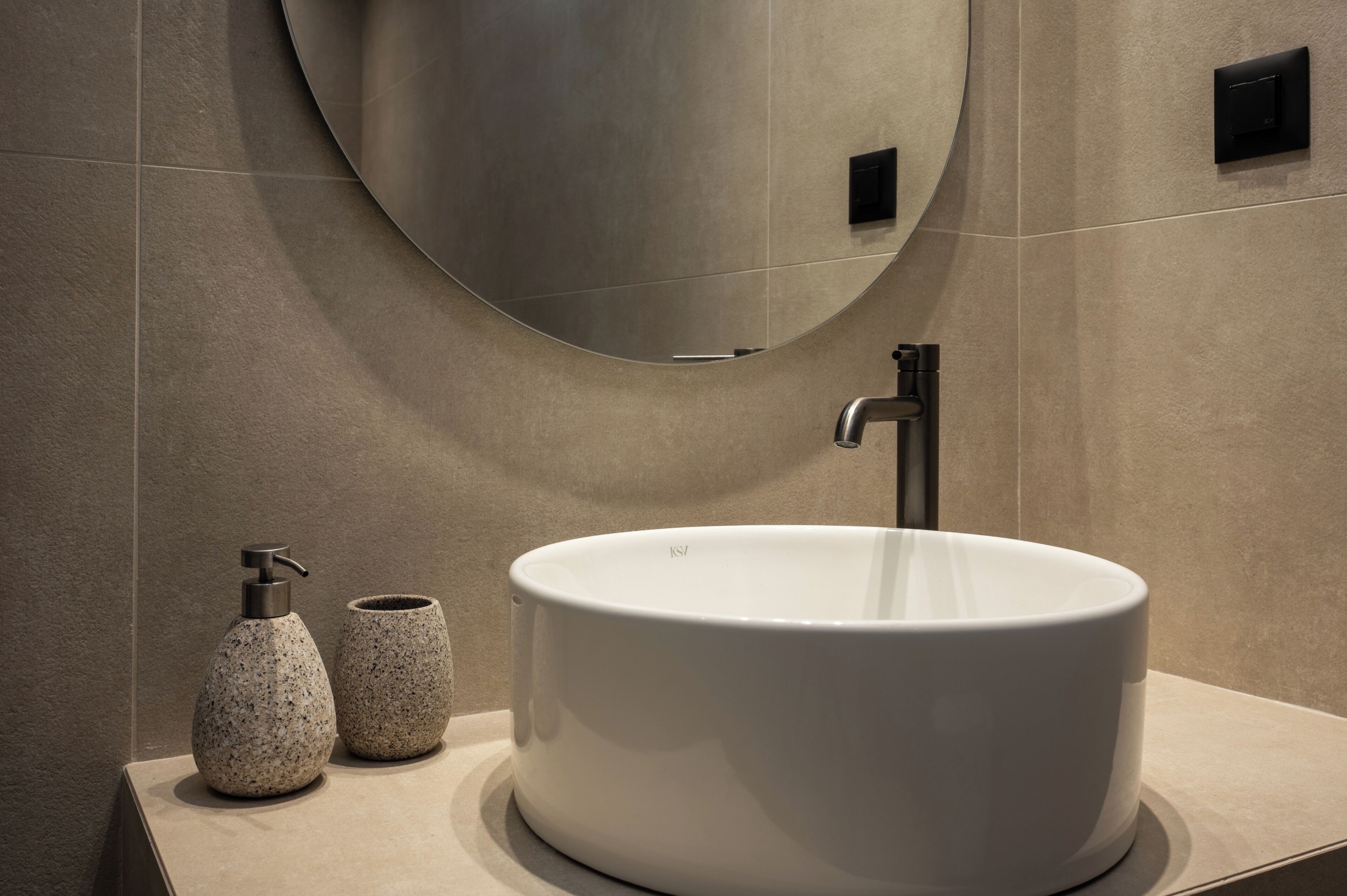 A modern bathroom with a large white sink, a round mirror, and two speckled vases on the countertop.