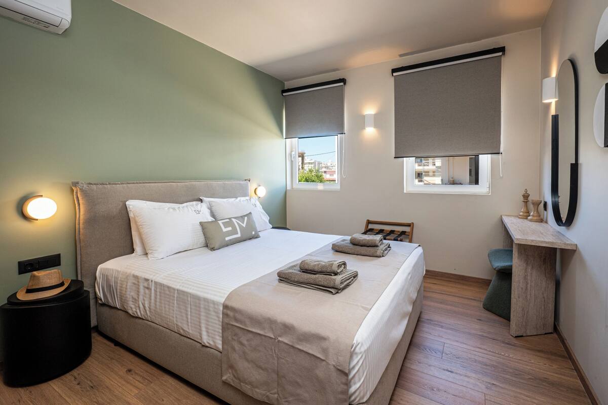 Ein ordentlich eingerichtetes Schlafzimmer mit Bett, Nachttischlampen, einem kleinen Tisch und einem Fenster mit Jalousien.