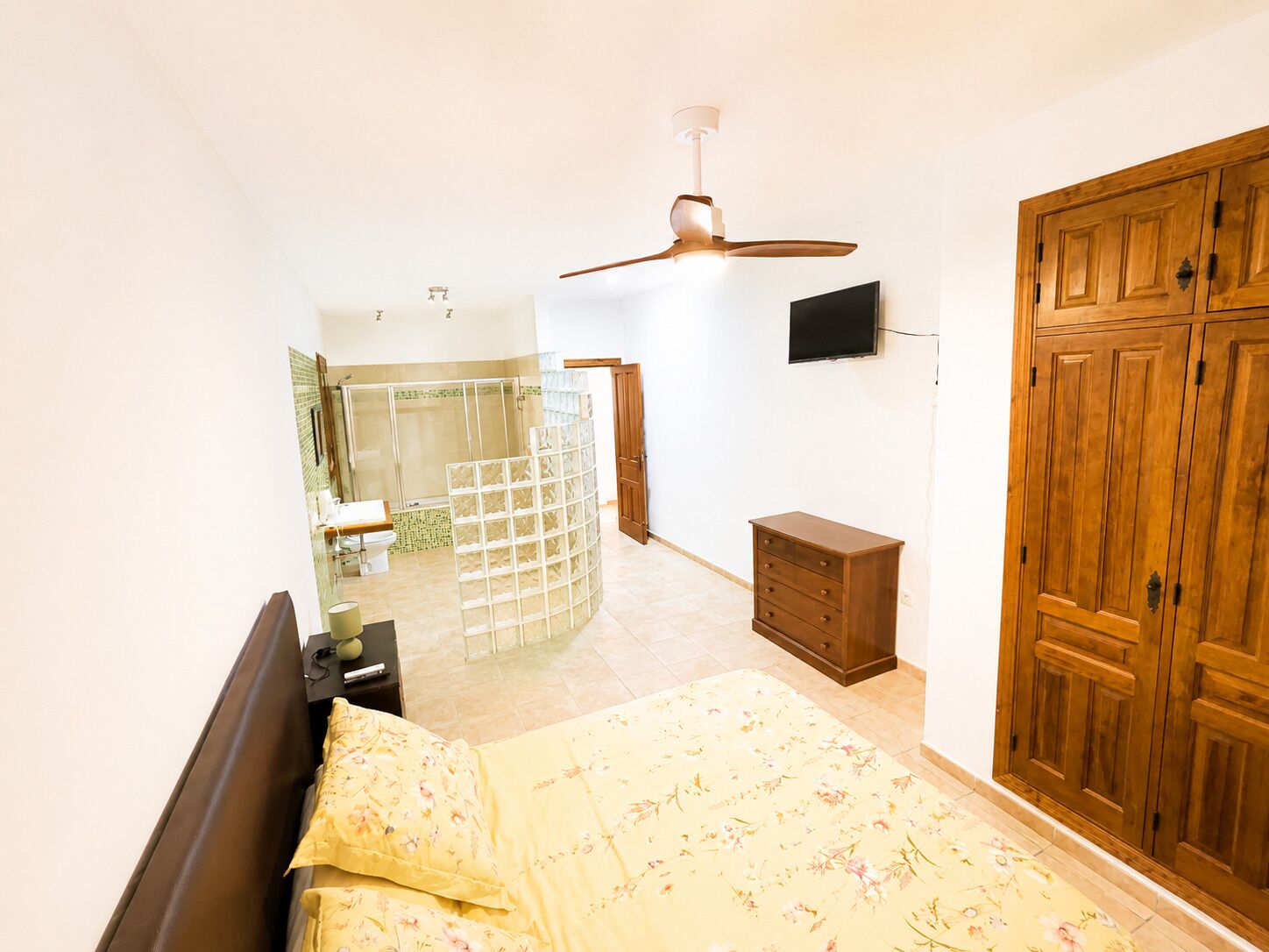 Un dormitorio con una cama, un armario de madera, un ventilador de techo, un televisor montado en la pared y un baño visible a través de un separador de vidrio.