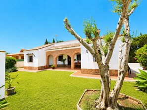 Ein zweigeschossiges Haus mit Terrasse, ein Baum im Vordergrund und ein klarer, blauer Himmel.