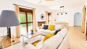Ein geräumiges Wohnzimmer mit einer Couch, einem Couchtisch und einem Fernseher. Es gibt einen Essbereich mit Tisch und Stühlen sowie einen Deckenventilator.