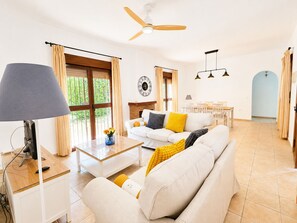 Ein geräumiges Wohnzimmer mit einer Couch, einem Couchtisch und einem Fernseher. Es gibt einen Essbereich mit Tisch und Stühlen sowie einen Deckenventilator.