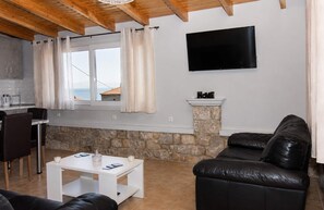 Un espacio habitable con una pared de piedra, un televisor de pantalla plana montado en la pared, un sofá de cuero negro, una mesa de centro blanca y vistas al mar a través de una ventana con cortinas.