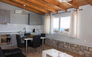 A modern kitchen with a stone wall, a dining area with a table and chairs, and a view of the sea through a window. - Emilia's Guest House with Sea View Balcony, Stoupa (Stoupa)