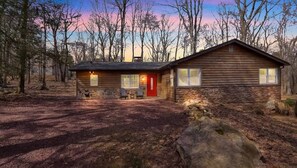A wooden cabin with a stone foundation, a red door, and a small porch with two chairs. - Rustic Woodland Cabin | Hot Tub | Cozy Escape (Lake Harmony)