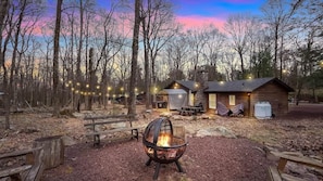 A rustic cabin with a fire pit, wooden benches, and string lights in a forest setting. - Rustic Woodland Cabin | Hot Tub | Cozy Escape (Lake Harmony)