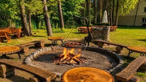 A circular fire pit surrounded by wooden benches and tables in a grassy outdoor area with trees and a building in the background. - Apartment with swimming pool, Lukecin (Łukęcin)