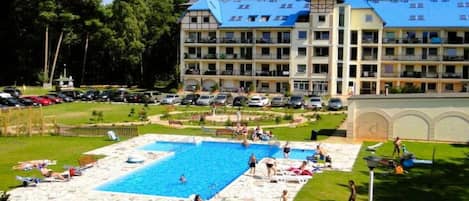 A hotel with a swimming pool surrounded by a landscaped garden, multiple balconies, and a parking area.