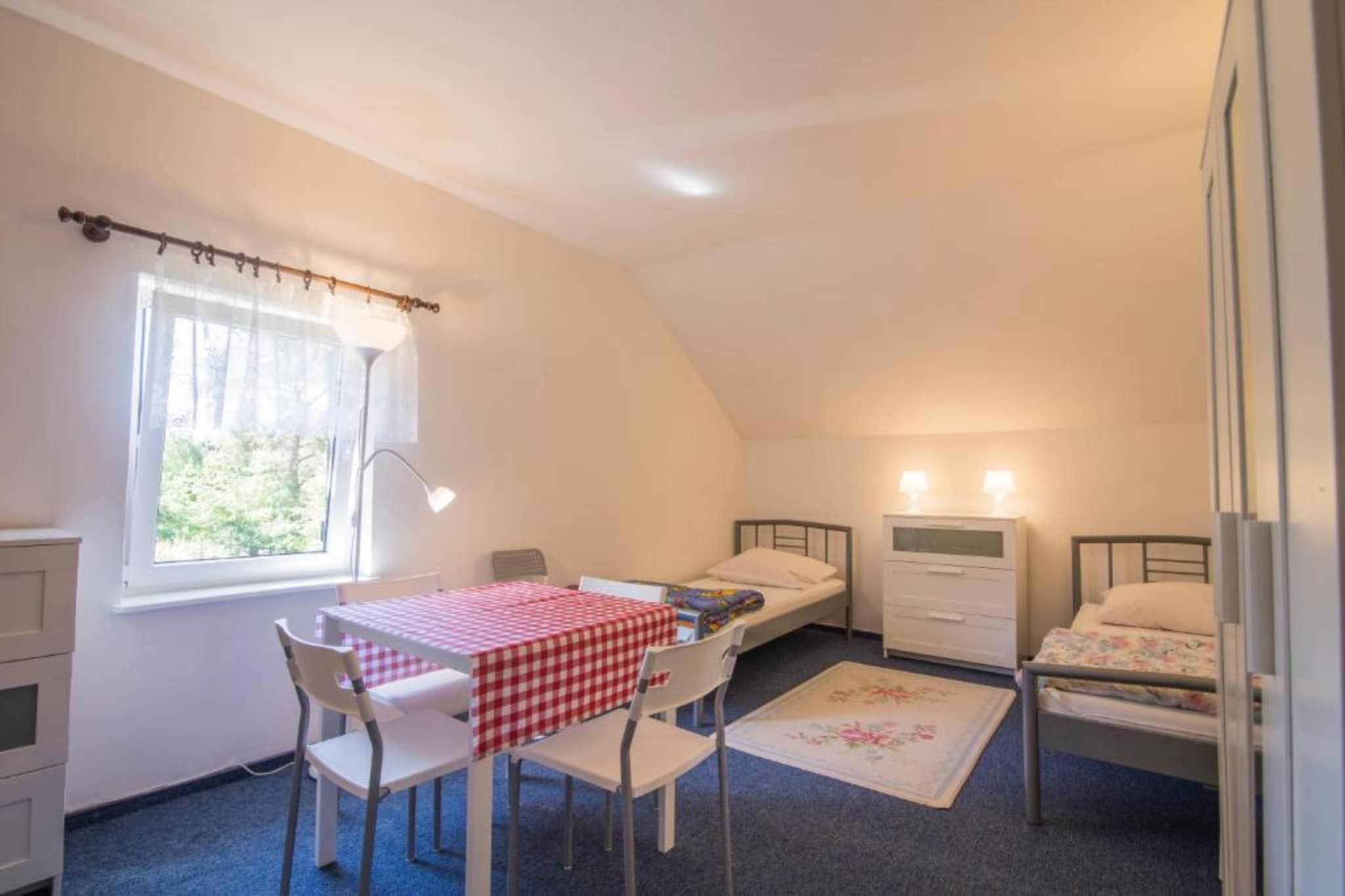 Quarto com duas camas de solteiro, uma mesinha com toalha xadrez, uma cadeira, uma janela com cortinas e um abajur.