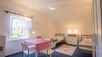 Quarto com duas camas de solteiro, uma mesinha com toalha xadrez, uma cadeira, uma janela com cortinas e um abajur.
