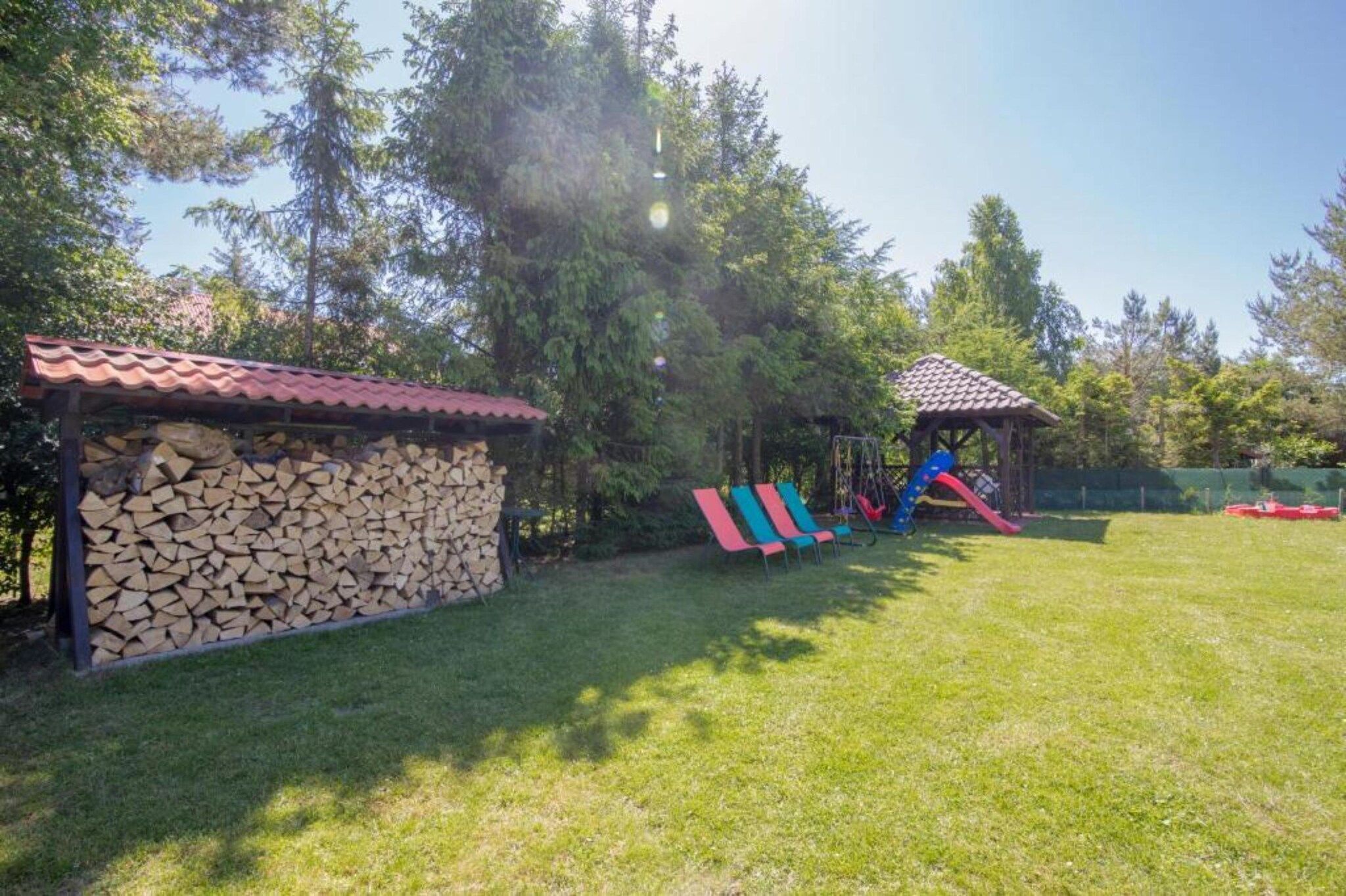 A grassy outdoor area with a playground featuring slides and swings, a covered wooden structure, and a stack of firewood.