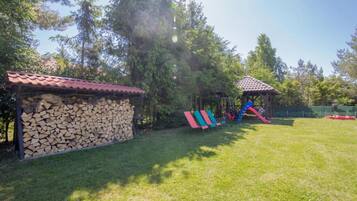 A grassy outdoor area with a playground featuring slides and swings, a covered wooden structure, and a stack of firewood.
