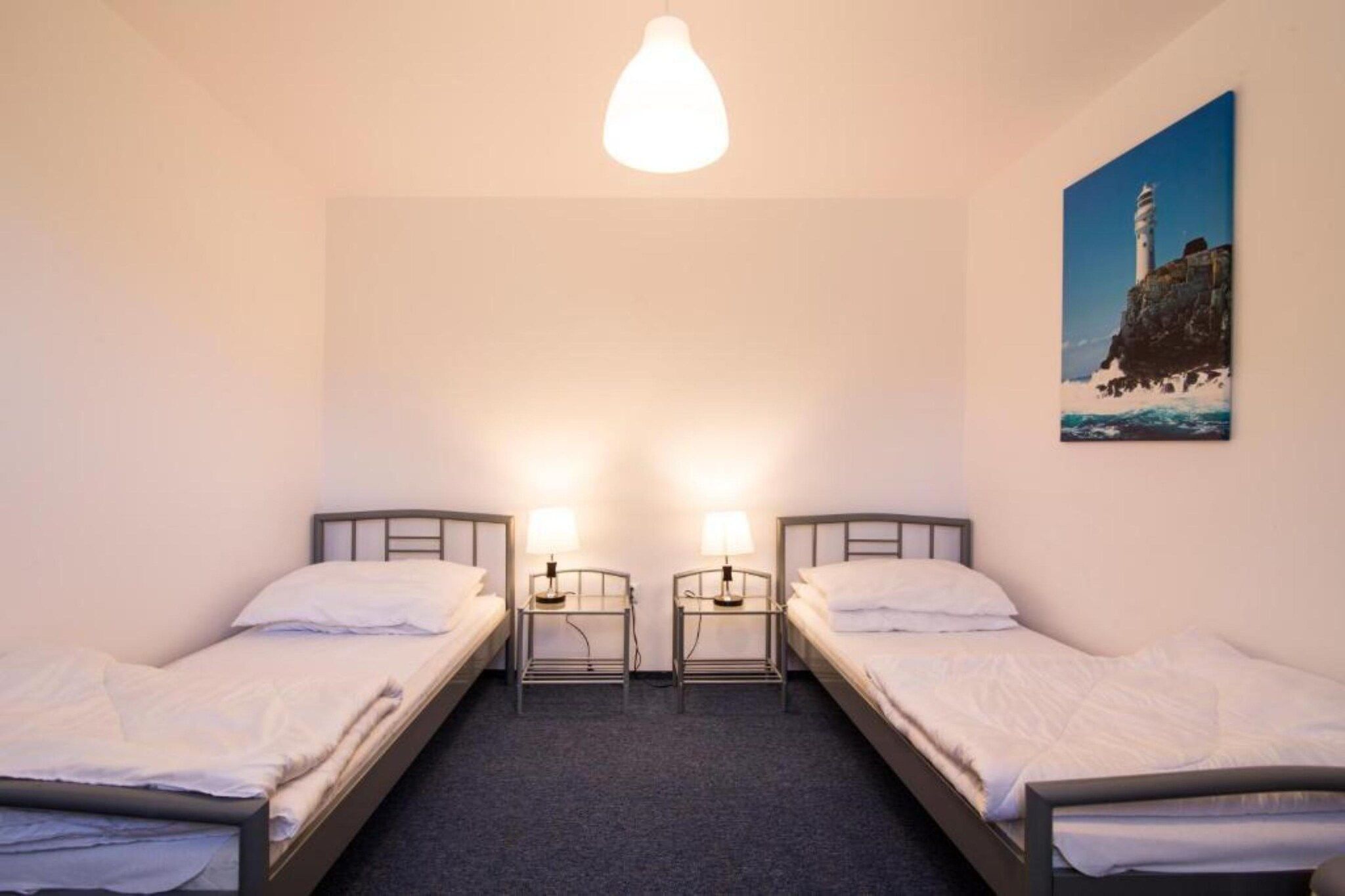 A room with two single beds, a light fixture above, and a painting of a lighthouse on the wall.