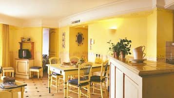 Un comedor con una mesa y sillas amarillas, un pequeño rincón de cocina con un refrigerador, y una planta en maceta sobre la encimera.
