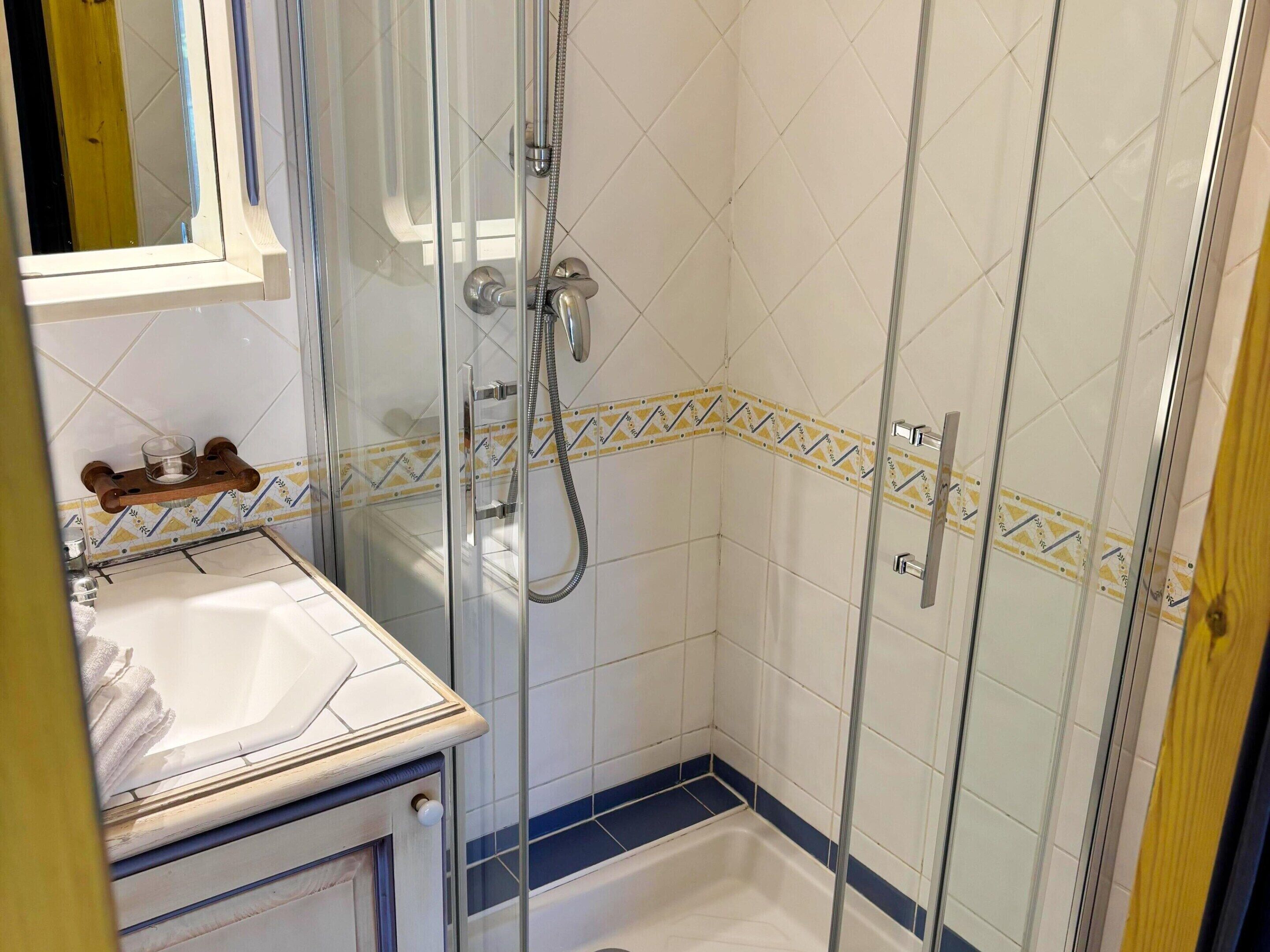 Eine begehbare Dusche mit Glaswänden, einem Handbrausekopf, einer kleinen Ablage mit einem Glasbehälter sowie ein weißes Waschbecken mit Handtuch.