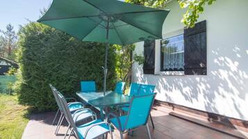 An outdoor patio with a table and chairs, a green umbrella, and a white building with a window.