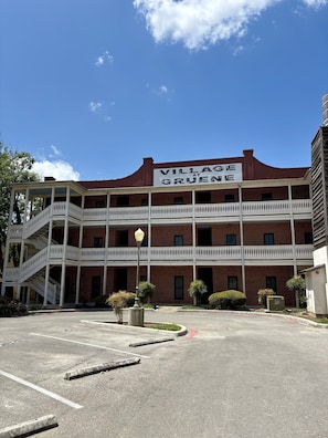 Un edificio de dos pisos con un cartel que dice 'Village at Grune'. Tiene fachada de ladrillo, barandales blancos y un estacionamiento al frente.