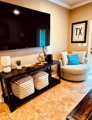 A living room with a flat-screen TV mounted on the wall, a dark wooden console table with decorative items, a beige armchair with a blue pillow, and a framed 'TX' sign on the wall.
