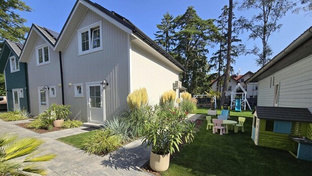 Ett bostadsområde med ett hus som har en veranda, en liten trädgård med olika växter och en lekplats för barn med en rutschkana och gungor.