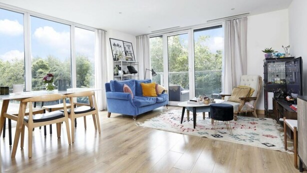 A spacious living room with a large window, a blue sofa, wooden chairs, a small round table, and a cabinet with glass doors. - Luxury Flat in Chelsea Bridge (Greater London)