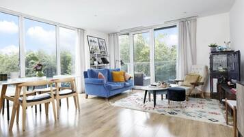 A spacious living room with a large window, a blue sofa, wooden chairs, a small round table, and a cabinet with glass doors.
