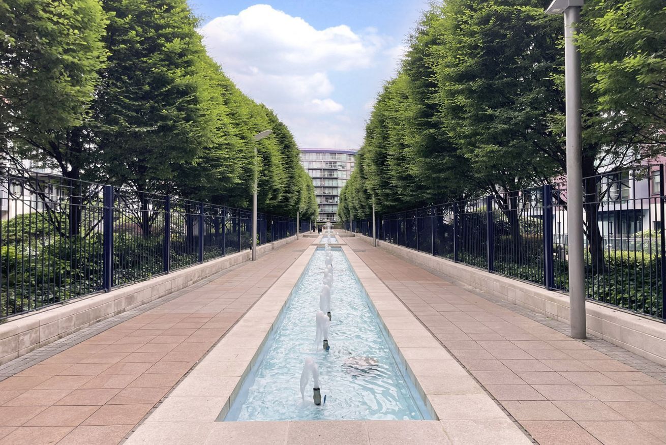 A long, straight pathway with a central water feature, flanked by trees and metal fences.