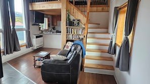 A cozy living room with a leather sofa, a bookshelf, a staircase, and a kitchen area.