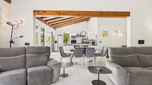 Una sala moderna con sofás grises, una mesa de comedor con sillas, una cocina al fondo y una viga de techo de madera.